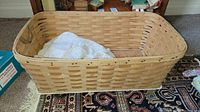 Photo of a rectangular vintage woven wooden baby bassinet containing a white lace-trimmed baby blanket and some baby clothes inside, placed on a patterned rug.