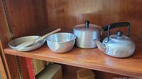 Photo showing the full set of vintage child's kitchen play items including frying pan, mixing bowl, saucepan, teapot, and wooden spoon on a wooden shelf.
