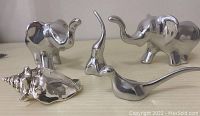 Four stainless steel sculptures including two elephants, one abstract shell, and one bird, plus another abstract form all polished and shiny.