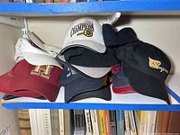 Six baseball caps stacked on a shelf, showing different team logos and designs.