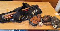 Photo showing three baseball gloves, two baseball bats in a Louisville travel tote bag, and one baseball on a wooden table.