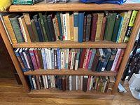 Three shelves of assorted hardback and paperback books arranged in wooden bookcases showcasing a variety of titles and conditions