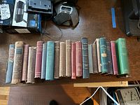 Row of 18 vintage hardback books arranged on a wooden table, showing spine titles and colors including various earth tones and blues.