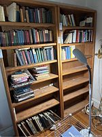 View of two oak bookcases with multiple shelves filled with assorted books including hardbacks and paperbacks, with some books stacked horizontally.