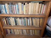 Three wooden shelves filled with a variety of books, mostly paperback with some hardcovers, showing wear and discoloration consistent with age. Books are tightly packed and span several decades of publication.