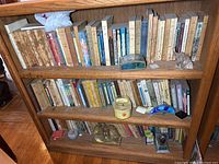 Three shelves filled with a mix of old hardback and paperback books, worn condition. Decorative items include a glass eagle bookend, a small clear animal figurine, a candle, and a small metal box. Books are arranged upright with some leaning.