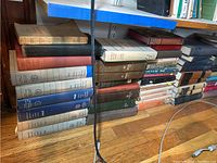 Stack of hardback and paperback books organized in piles on the floor beneath a bookshelf, showing titles and spines mostly worn.