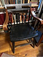 Wood arm chair with black painted seat and spindles, natural wood armrests, engraved emblem on top backrest, turned legs and stretchers, photographed with books in background.