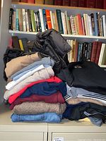 Stack of folded clothing items including sweaters, jackets, and jeans placed on a surface with books in the background