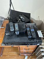 Panasonic DVD/VHS combo player and Samsung Blu-ray player with four remote controls placed on wooden desk, cables behind.