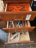 Three wooden drawers with brass handles partially open, showing kitchen utensils inside the drawers with an orange cutlery organizer in top drawer.