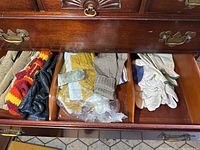 Top drawer with multiple pairs of gloves and mittens including knit mittens, black leather gloves, some yellow knit mittens in a plastic bag, and white work gloves.