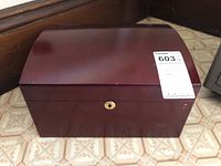 Front view of dark cherry wooden humidor box with curved lid and gold-tone lock, placed on tile floor.