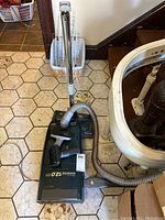 Photo showing full vacuum cleaner including chrome handle, hose attached to the dark green canister body, on tiled floor.