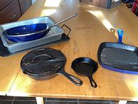 Full view of lot items on wooden table including Lodge cast iron skillets, Cuisinart grill pan, blue frying pan, and baking dishes