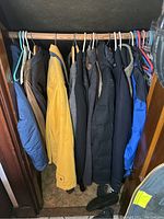 View of seven men's coats and jackets hanging on a wooden rod in a closet, showing variety of colors and styles.