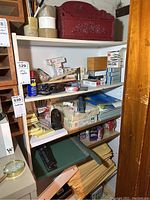 White shelving unit filled with various office supplies including tape, clips, pens, and a red wood organizer box on top.