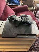 PlayStation 3 console with three wired controllers on top, dusty surface visible.