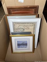 Box containing five framed artworks and plaque, varied frame styles and sizes visible from top angle