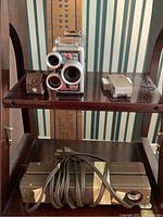 Front view of the vintage Bell & Howell 8mm movie camera and two small metal accessories on a shelf above two metal lighting bars with power cords.