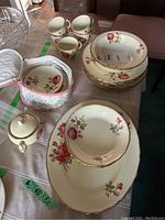 Photo of multiple china items including plates, tea cups, sugar bowl, and saucers with 22kt gold trim and rose floral pattern laid out on a table.