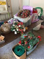 Multiple tubs on a table containing various Christmas decorations including artificial floral arrangements and large light globe.