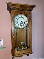 Front view of wall clock showing wooden case with glass panel, Roman numeral dial, and metal pendulum visible inside.