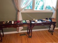 Wide view of two wooden side tables with assorted costume jewelry, including necklaces, brooches, cufflinks, wristwatch, and jewelry boxes, near window with curtains.