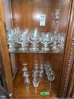 Glassware set inside wooden cabinet shelf including brandy glasses, smaller liqueur glasses, and decanter