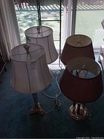 Four table lamps in two pairs placed near a sliding glass door with natural light. Two lamps with white fabric shades and silver-tone tripod bases, two lamps with brown pleated shades and brass bases.