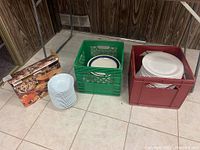 Two crates with stacked plates and bowls on tiled floor, additional boxed item and stacked small plates.
