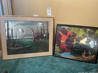 Two framed prints standing against a wall showing detailed scenic artworks: one with birds over swamp, the other with watermill and children.