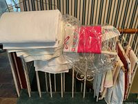 Assortment of mostly white tablecloths hanging and folded over a railing. Some have floral embroidered borders with red and green colors. Some are still in plastic wrapping.