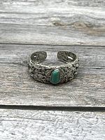 Front-facing photo of silver-tone bangle bracelet with floral engraving and turquoise inset stone on wooden background.