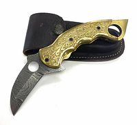 Folded hunting knife with gold-colored engraved handle resting on stitched leather pouch