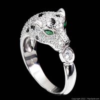 Angle shot showing the detailed cat head with green crystal eyes, black spots, and clear crystals, holding a round clear crystal in mouth on silver-tone band.