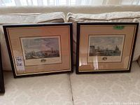 Two framed antique French port prints displayed on a couch, showing full framed presentation with beige mats and dark wood frames.