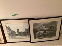 Photo showing two framed black and white vintage-style drawings leaning against a wall. One drawing depicts a historic cityscape with a tower; the other a mining industrial building.
