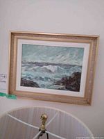 Framed oil painting showing a seascape with ocean waves and rocks. The painting is mounted on a wall above a lamp.