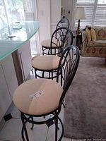 Photo showing three bar stools arranged in a row at a kitchen counter, featuring black metal frames with arched backrests and beige padded round seats.