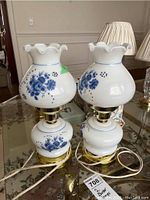 Pair of matching glass boudoir lamps on reflective glass table showing white glass shades with blue floral patterns, brass bases, and white cords.