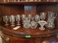 Crystal glassware set displayed on wooden cabinet shelf including stemware glasses, a pitcher, covered relish jars and nut dish.