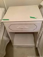 Photo of the night table showing the front drawer with decorative painted carving and open shelf below, cream colored wood painted finish.