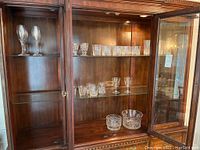 Two shelves inside wooden cabinet displaying crystal tumblers and stemware including wine and sherry glasses plus two glass serving bowls on bottom shelf.