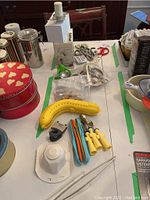 Overview of the lot showing various kitchen tools and accessories including kids utensils, banana keeper, tongs, scissors, Cuisinart attachments, and more arranged on a table.