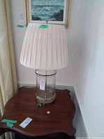 Full view of table lamp with clear glass base and pleated fabric shade on a wooden table, set against a wall with artwork.