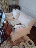 Photo showing white upholstered love seat with cushions, placed on patterned rug, surrounded by framed artwork and furniture.