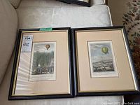 Both framed vintage French air balloon prints side by side on a cream sofa, showing full frames and mats.