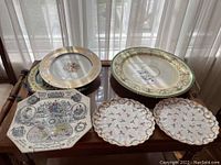 Photo showing multiple decorative plates on a wooden table including an octagonal commemorative plate, large gold and green border plates, and two small floral scalloped-edge plates.