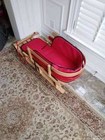 Wooden sleigh with red liner from an angled top view showing full length of the sleigh resting on a patterned rug indoors.